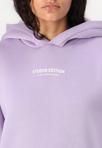 Lilla hettegensare laget av mykt stoff med en stor hette. Har "STUDIO EDITION" og "ALPHA INDUSTRIES INC." trykt i hvitt på forsiden.