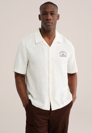 Mężczyzna w krótkim, białym koszuli na guziki z napisem „Sunset State Clubhouse”, z rękami w kieszeniach, stoi na beżowym tle.