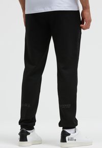 Schwarze Jogginghosen aus Baumwolle mit schmaler Passform, seitlicher Markenkennzeichnung, Bündchen und Pflegesymbolen in der Nähe des Knöchels. Kombiniert mit weißen Sneakers.