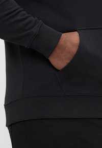 Czarna bluza z kapturem z prążkowanymi mankietami i kieszenią typu kangur. Wykonana z gładkiego materiału o matowym wykończeniu. Dolny brzeg lekko zaokrąglony.