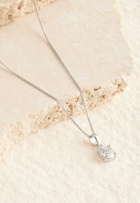 Silberne Halskette mit rundem, klarem Anhänger in Zargenfassung, auf strukturiertem beigem Stein-Hintergrund platziert.