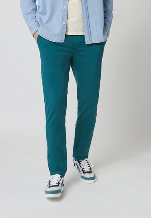 Man met turquoise broek, witte en turquoise sneakers, lichtblauw overhemd met knopen, staat met de handen in de zakken op een effen lichte achtergrond.