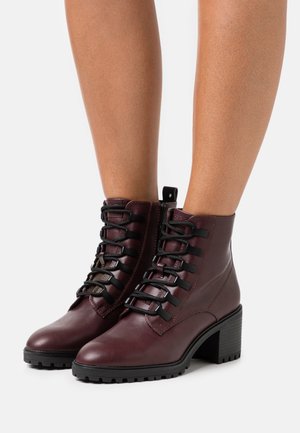 Bottes en cuir bordeaux montant à la cheville, avec des lacets noirs, des talons blocs épais et une semelle crantée, portées sur des jambes nues.