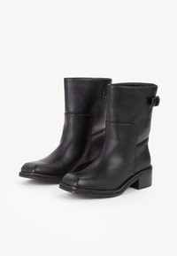 Para czarnych skórzanych botków do połowy łydki z niskimi słupkowymi obcasami i małą klamrą przy górze na białym tle.