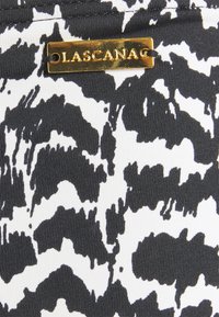 Schwarz-weißer, gemusterter Stoff mit kräftigen, abstrakten Formen. Enthält ein goldenes "LASCANAG"-Logoetikett, das die Textur und das Designdetail hervorhebt.