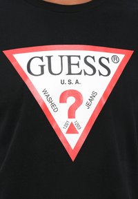 Tricou negru din bumbac, prezentând un triunghi mare alb cu accente roșii. "GUESS" și "WASHED JEANS" imprimate vizibil, cu un design de semn de întrebare.