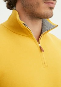 Homme portant un pull zippé jaune moutarde avec un col intérieur gris, montrant la partie inférieure du visage et le cou.