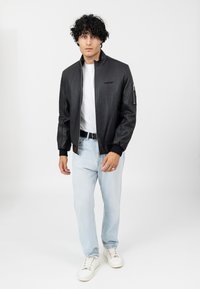 Giubbotto bomber in pelle nera con colletto alto, chiusura con zip, tasche laterali, abbinato a una t-shirt bianca e jeans azzurri chiari, indossato con sneakers bianche.