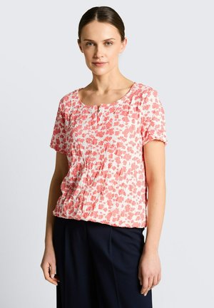 Femme portant un chemisier à manches courtes avec des motifs floraux roses et blancs, col rond, et un pantalon noir, sur un fond uni clair.