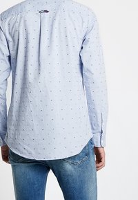 Lichtblauw overhemd met kleine marineblauwe stippen, lange mouwen en een plooi aan de achterkant. Gecombineerd met blauwe denimjeans.