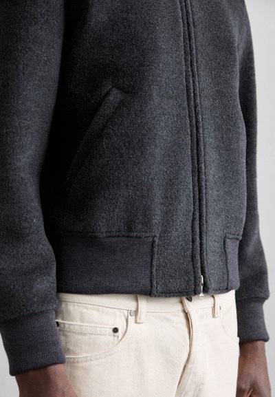 Chaqueta de forro polar gris oscuro con cierre de cremallera, puños acanalados y bolsillos laterales, combinada con pantalones beige claros. Textura suave y corte casual.