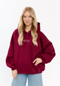 Felpa oversize bordeaux realizzata in tessuto morbido, dotata di cappuccio con cordino e polsini elasticizzati, con un discreto dettaglio logo sul davanti.