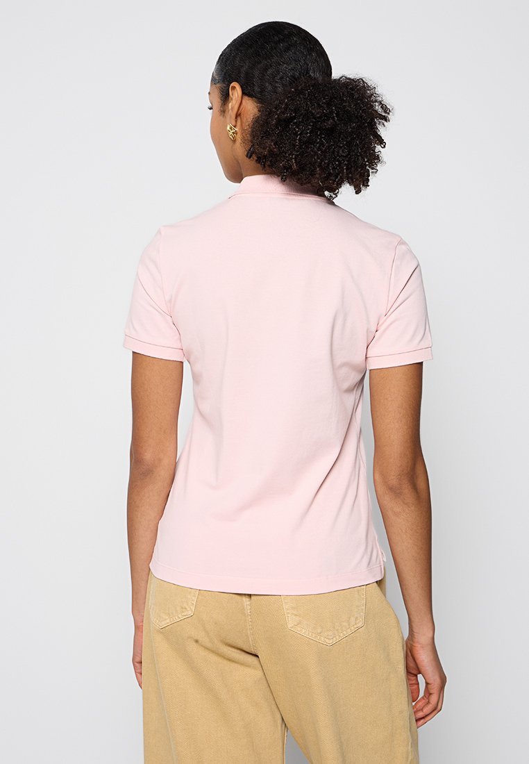 Femme aux cheveux bouclés en chignon bas, portant un polo rose clair à manches courtes et un pantalon beige, vue de dos.