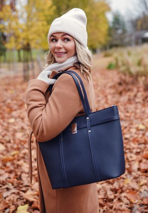 Marineblaue Tragetasche aus glattem Material mit zwei Griffen, Metallakzenten und einer strukturierten Form. Kombiniert mit einem beigen Mantel und einem weißen Schal.