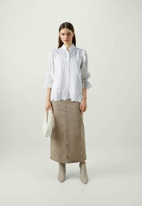 Blusa branca de botão com mangas franzidas, combinada com uma saia de denim bege e botins cinza claro. Modelo segurando uma mala branca.