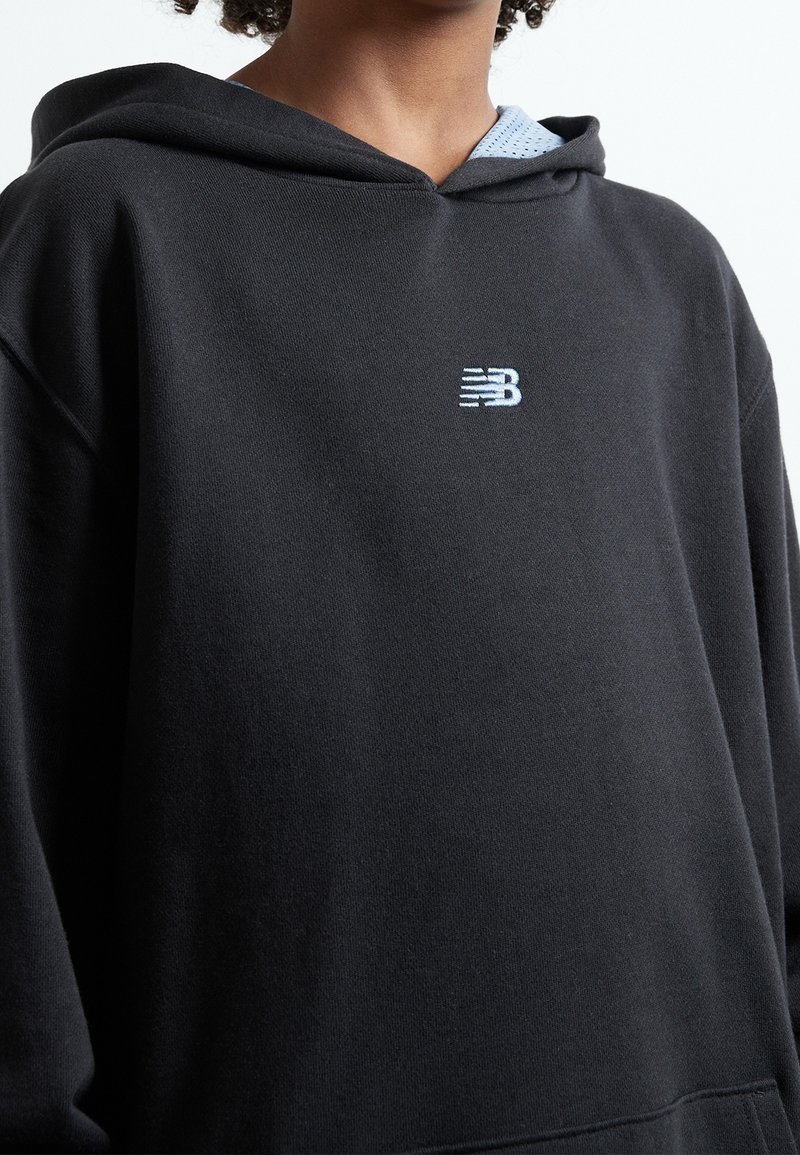 Persona che indossa un hoodie nero con un piccolo logo bianco di New Balance centrato sul petto, tessuto blu chiaro visibile all'interno del cappuccio.