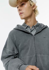 Graue Zip-Hoodie mit strukturierter Oberfläche, Kapuze und kleinem Logo. Darunter ein Button-Up-Hemd in einem passenden Grauton.