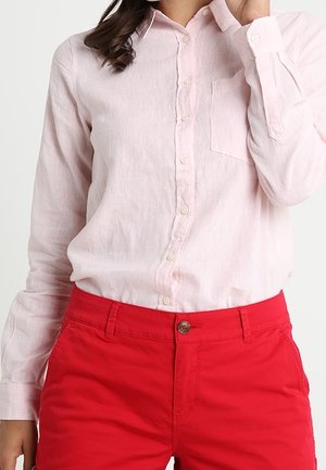 Kobieta ubrana w jasnoróżową koszulę z długim rękawem zapinaną na guziki, włożoną do jaskrawoczerwonych spodni z zapięciem na guzik i zamek.