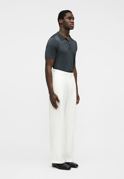 Muž stojící, oblečený v tmavě šedé polo košili s krátkým rukávem, bílých širokých kalhotách a černých botách, na jednobarevném světlém pozadí.