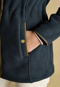 Námořnická fleecová bunda s kontrastním olivovým lemováním, boční kapsou so zlatým cvokom a texturovanou tkaninou. Teplý a ležérny dizajn.