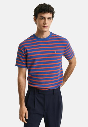 Blue and red striped short-sleeve t-shirt with a round neckline and a small logo detail on the chest. Worn with dark trousers.