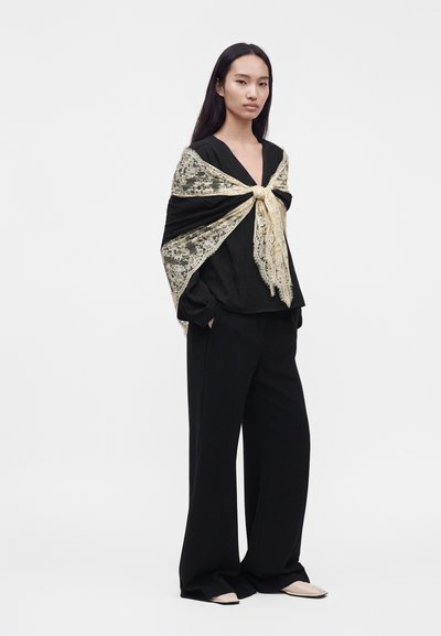 Femme aux longs cheveux foncés portant une tenue noire ample et un grand châle en dentelle beige noué en nœud papillon sur ses épaules.