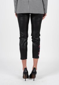 Pantalons en cuir noir coupés avec un design ajusté, présentant des broderies florales multicolores sur l'ourlet droit. Portés avec des chaussures à talons hauts noires.