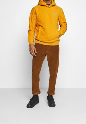Sudadera amarilla con capucha y logo, combinada con pantalones de pana marrón y zapatillas negras. Presenta un corte relajado y una tela texturizada.