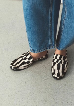 Stopy w wsuwanych mokasynach w paski zebry, połączonych z niebieskimi, szerokimi spodniami o długości do łydki, stojące na jasnoszarej podłodze.