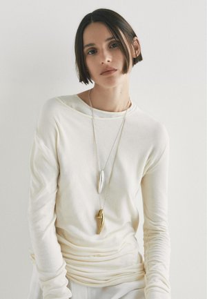 Kremowa koszula z długim rękawem o luźnym kroju i podwiniętym hemie. Posiada nałożone metaliczne naszyjniki - jeden srebrny, drugi złoty - na gładkiej fakturze.