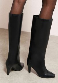 Cizme negre până la genunchi, din piele netedă, cu vârf ascuțit și toc subțire stiletto. Au o siluetă elegantă și ajustată.