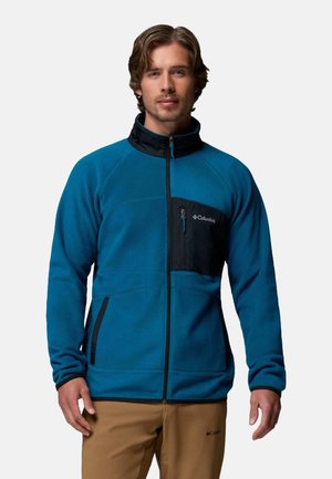 Mann trägt eine blaue Columbia Fleecejacke mit schwarzen Reißverschlusstaschen und beiger Hose, steht vor einem schlichten hellen Hintergrund.