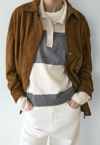 Brązowa zamszowa kurtka nałożona na kremowo-szary, paskowany sweter z prążkowanym dołem, zestawiona z białymi spodniami. Gładka tekstura na całej powierzchni.