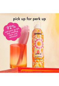 Shampoing sec Amika Perk Up dans un flacon vif et à motifs à côté d'un peigne en plastique rose dans un gobelet dégradé orange sur une surface plate rouge.