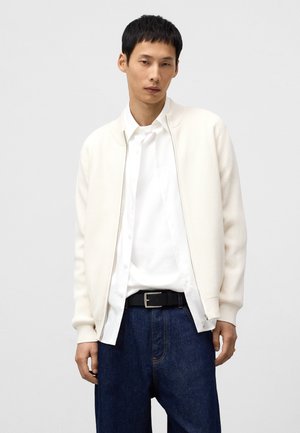 Knit, cream bomber jacket with a ribbed texture, zip closure, worn over a white shirt and dark denim pants, accompanied by a black belt.