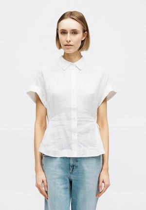 Γυναίκα με κοντά ανοιχτό καστανά μαλλιά που φοράει λευκό πουκάμισο με κοντά μανίκια και κουμπιά, καθώς και ανοιχτό μπλε τζιν, μπροστά σε απλό φόντο.