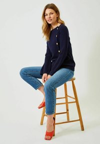 Marinblå tröja med guldfärgade knappar, lättstickad, matchad med ljusblå tajta jeans och röda öppna klackskor, sittande på en träpall.