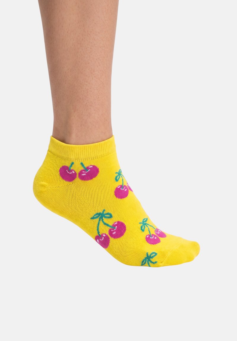 Calzino giallo alla caviglia con motivi di ciliegie rosa indossato su un piede su sfondo bianco.
