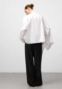 Donna con i capelli raccolti in uno chignon, che indossa una camicetta bianca ampia e pantaloni neri larghi, in piedi di spalle su uno sfondo grigio chiaro uniforme.