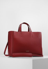 Sac tote en cuir rouge avec un design structuré, poignées doubles, bandoulière ajustable et logo Calvin Klein sur le devant. Texture lisse.