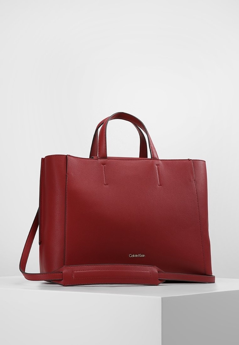 Sac tote en cuir rouge avec un design structuré, poignées doubles, bandoulière ajustable et logo Calvin Klein sur le devant. Texture lisse.