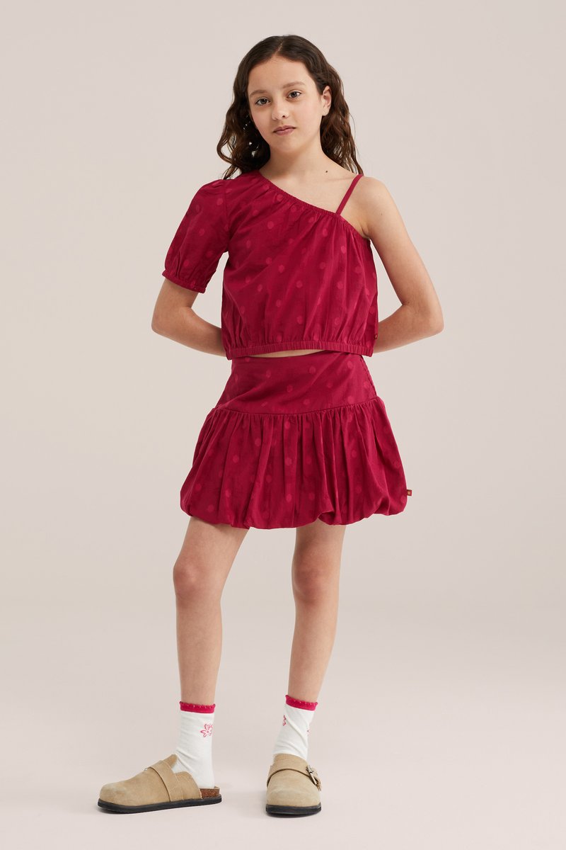 Fille portant un top rouge une épaule et une jupe assortie, avec des chaussettes blanches et des chaussures beiges sans lacets, debout les mains derrière le dos.
