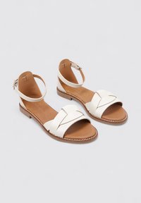 Sandales en cuir blanc avec semelle plate, présentant des brides croisées et un support de cheville. Semelle intérieure marron et détail de couture décoratif.