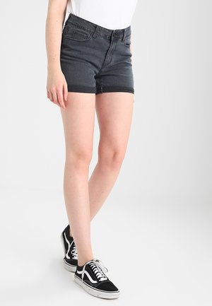 Person iført sorte denimshorts, hvid top og sorte sneakers, der står mod en ensartet lys baggrund.