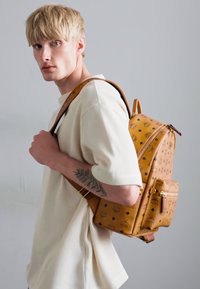 Sac à dos en cuir tan avec des accents à motifs, deux sangles, poche avant et quincaillerie dorée. Modèle portant un t-shirt blanc à côtes.