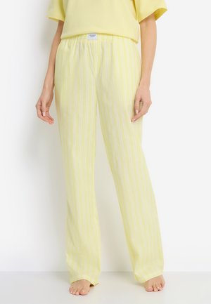 Frau in locker sitzender Hose mit gelb-weißen vertikalen Streifen und einem einfarbigen, hellgelben, kurzärmligen Oberteil, steht barfuß vor weißem Hintergrund.