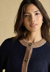 Mladá žena s dlhými tmavými vlasmi má na sebe námornícky sveter s béžovým rebrovaným golierom a zapínaním na gombíky, pózuje na béžovom pozadí.