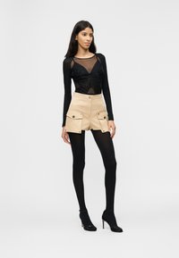 Γυναίκα που στέκεται φορώντας μαύρο διάφανο μακρυμάνικο τοπ, μπεζ σορτς ψηλόμεσο με τσέπες με καπάκια, μαύρες καλσόν και μαύρες ψηλοτάκουνες γόβες.