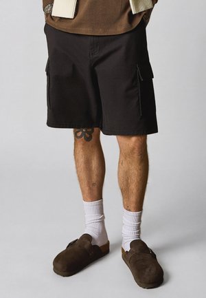 Férfi fekete cargo shortban, fehér zokniban és barna slip-on cipőben, lábán látható tetoválásokkal, egyszínű háttér előtt.