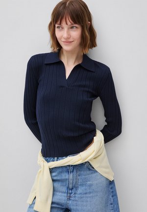 Vrouw met kort haar die een marineblauwe geribbelde longsleeve en lichtblauwe spijkerbroek draagt met een crème trui om de taille geknoopt.